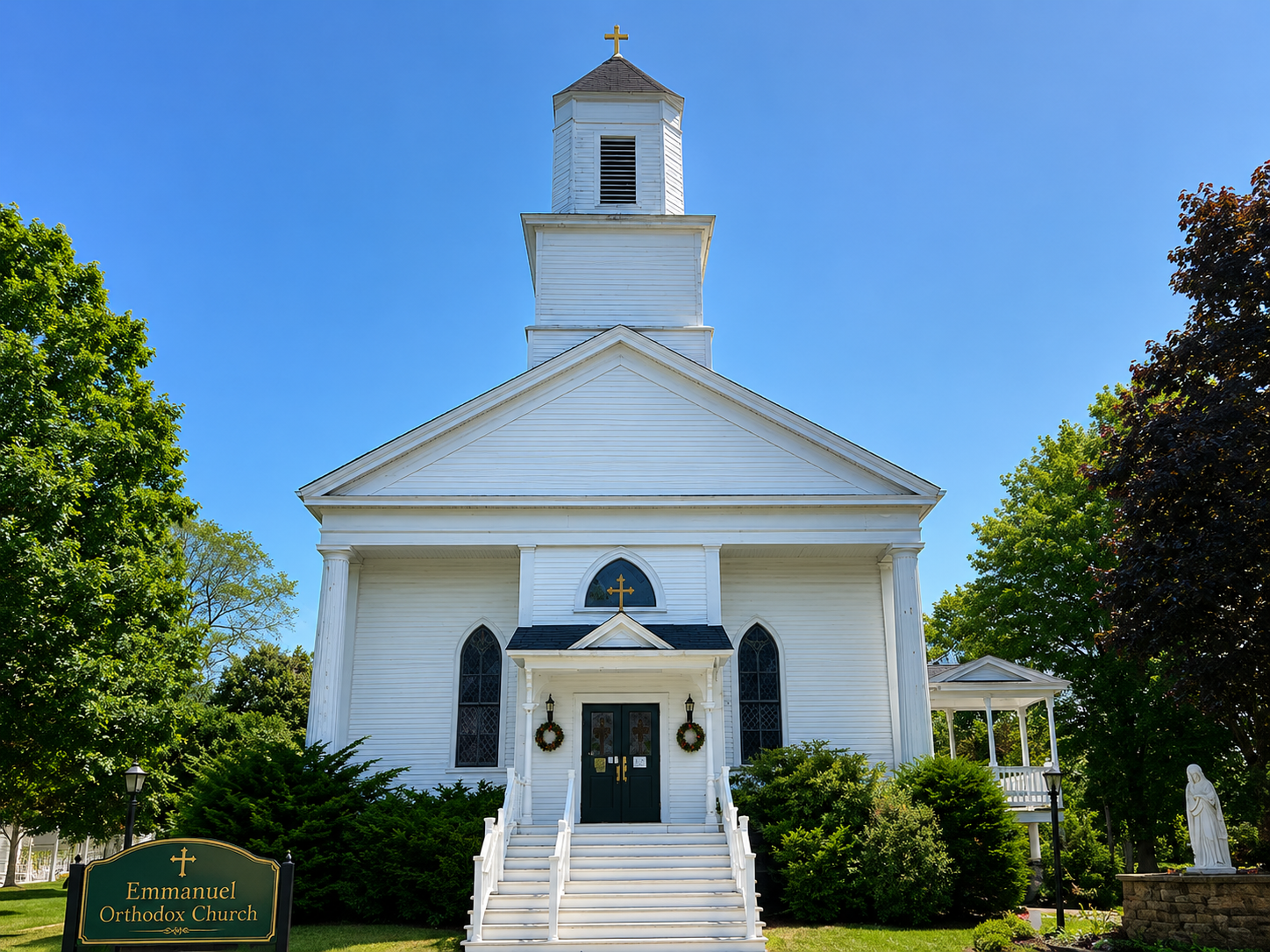The white clapboard building of Emmanuel Orthodox Church with its steeple and gold cross, set beneath a clear blue sky