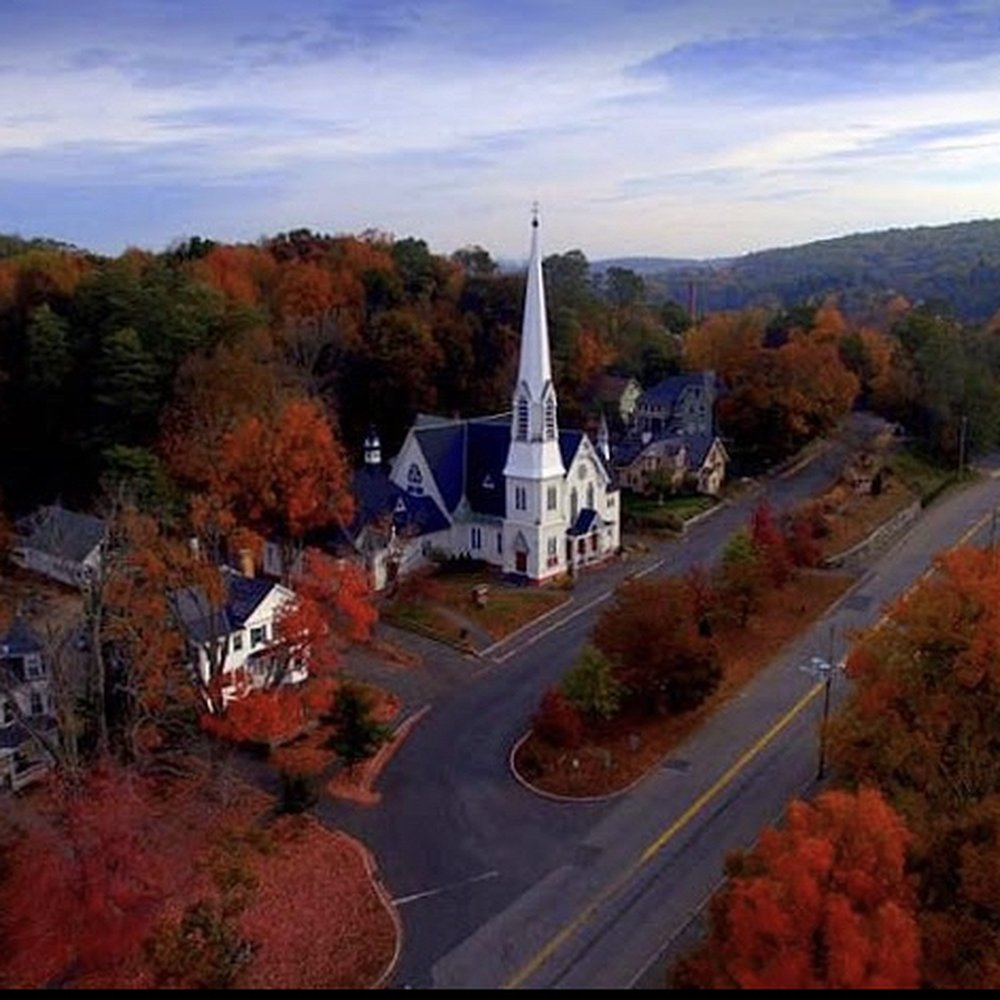 Aerial autumn view of the historic white-steepled Congregational church on the hill in the center of Warren, Massachusetts, surrounded by brilliant fall foliage — the parish's first permanent home.