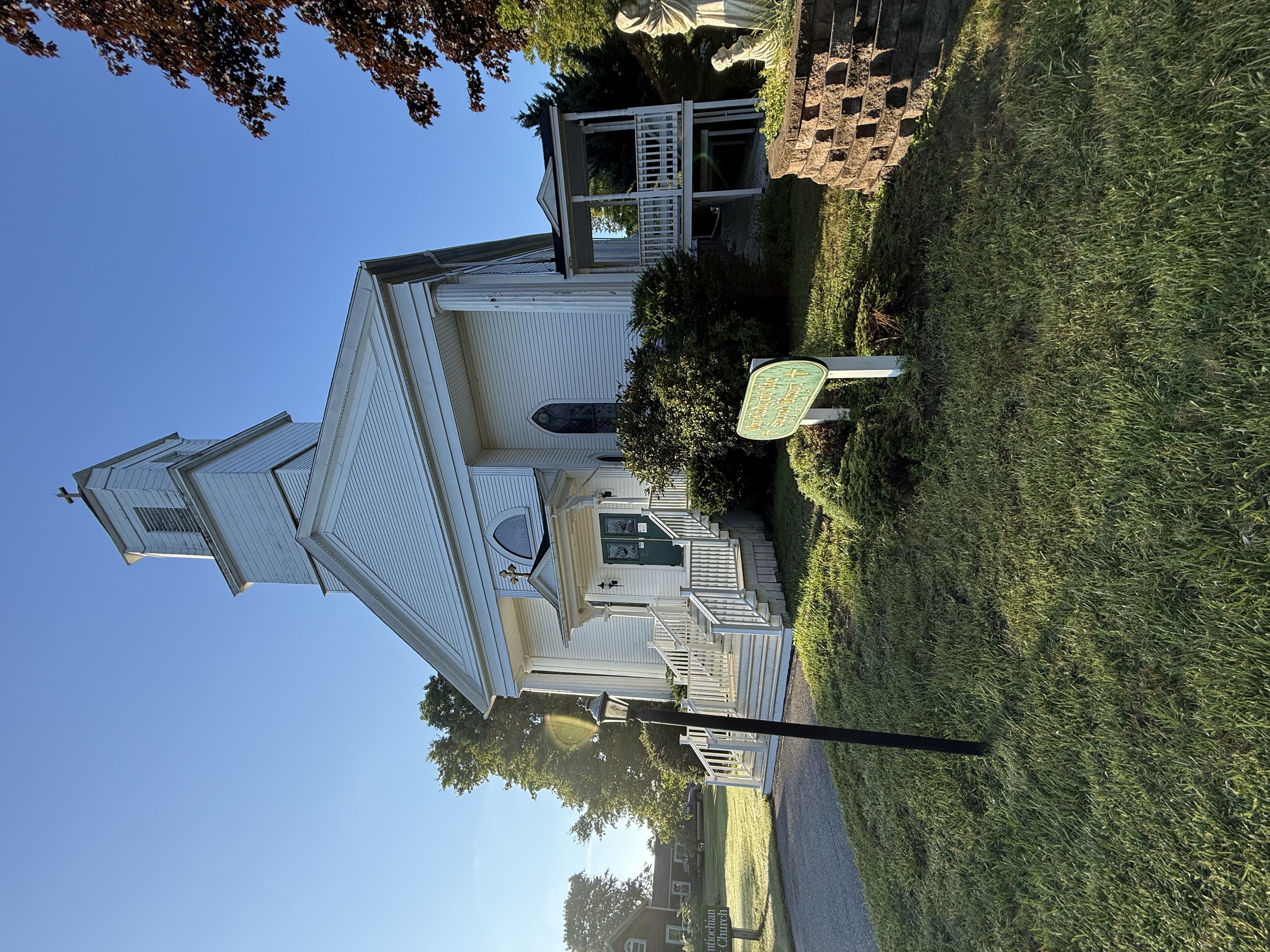 Full front view of the white clapboard church with its steeple and cross, portico, and parish sign in the morning sun