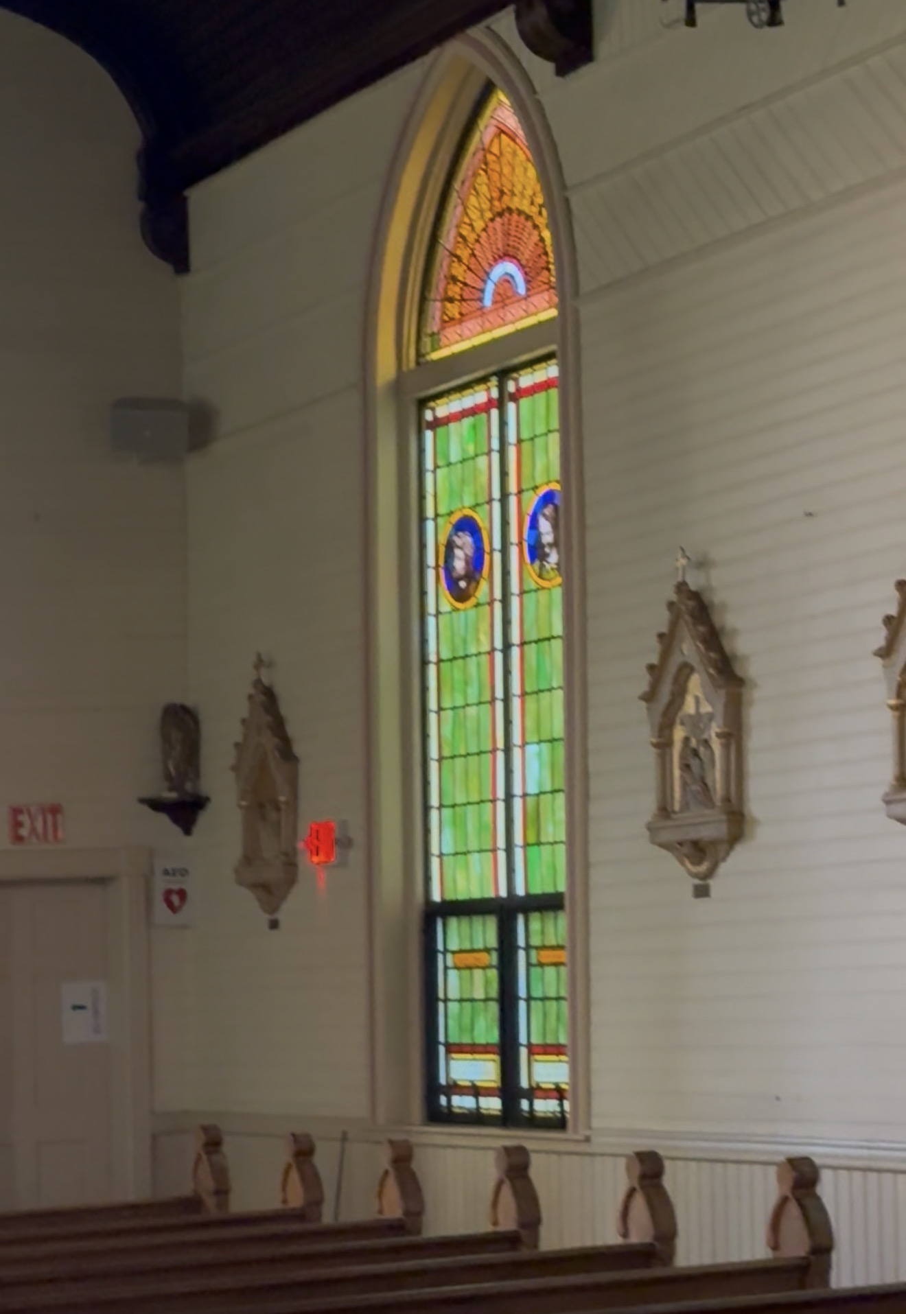 A tall Gothic stained glass window with green and amber glass, framed by carved Stations of the Cross on the clapboard wall